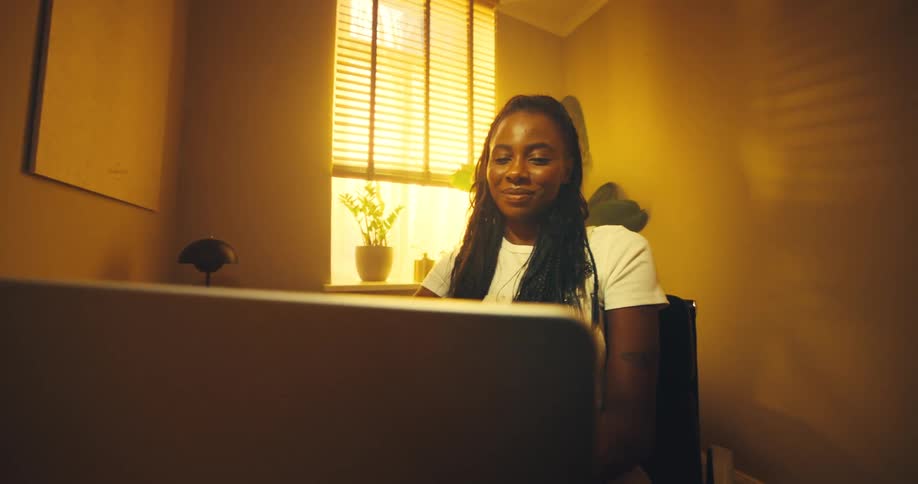 Sitting at her desk in a warmly lit room a woman opens her laptop and smiles her face illuminated as she prepares to begin her work