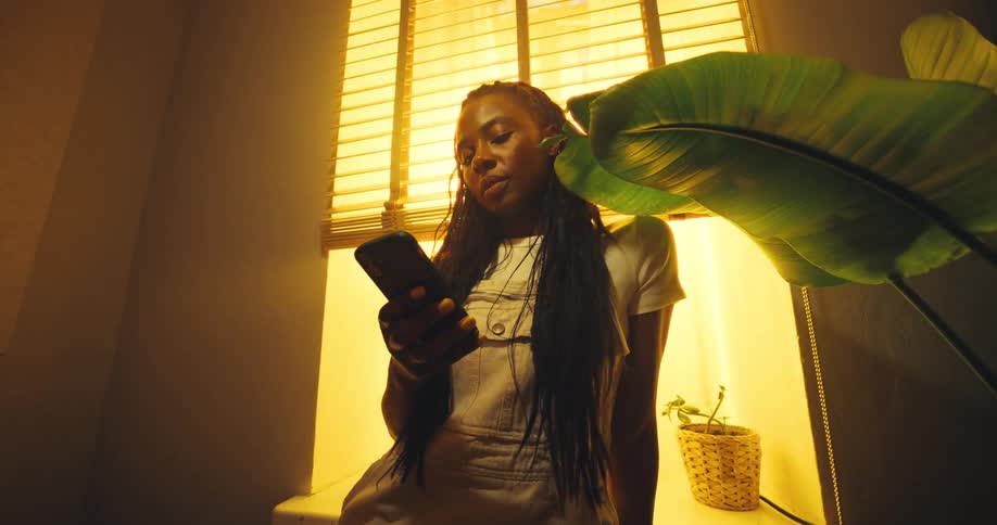 Young woman sitting near a window lifts her arm and scrolls though her phone