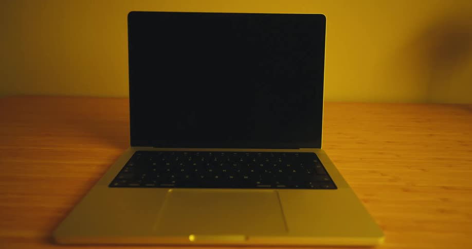 An open laptop with a dark blank screen sits centered on a wooden desk bathed in the warm yellow glow of an unseen light