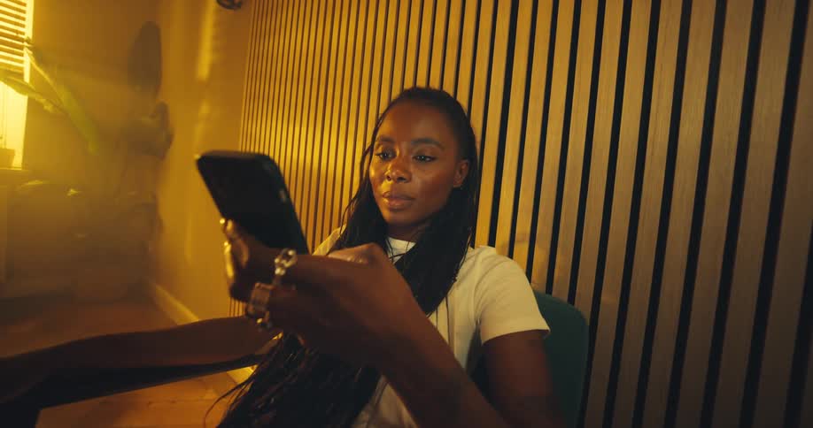 A woman is sitting in a chair in a room with yellow light picking up her cell phone and smiling as she looks at it