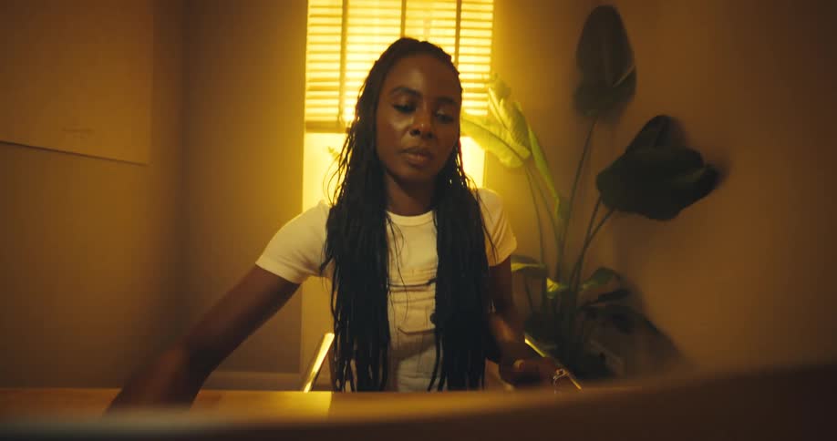 A young woman with long dark braids and a white t shirt sits at a wooden table in a room with warm lighting looking down