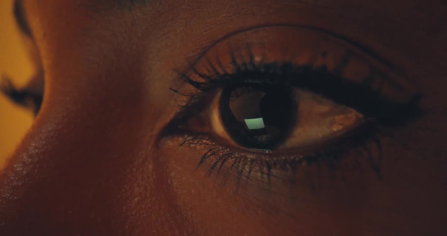 A tight extreme closeup focuses on a darkskinned eye with long dark eyelashes and thin eyeliner highly contrasted with a warm goldenorange background light