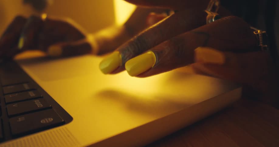 A woman gently taps her fingers on a laptop placed on a desk her fingers adorned with rings and her nails polished in yellow