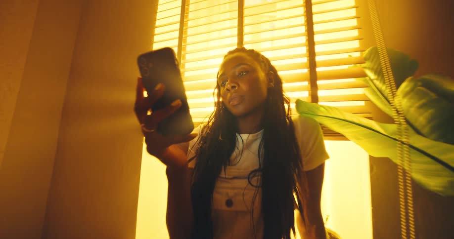 A woman stands by a window engrossed in her mobile phone in a warmly lit room