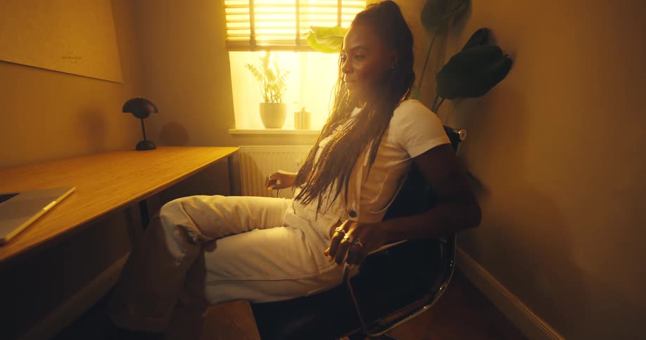 This video shows a woman with long braids and yellow nails transitioning from relaxing in her chair to actively typing on her laptop at a wooden deskthe scene is lit with a warm golden ambient light coming from a window with horizontal blinds creati