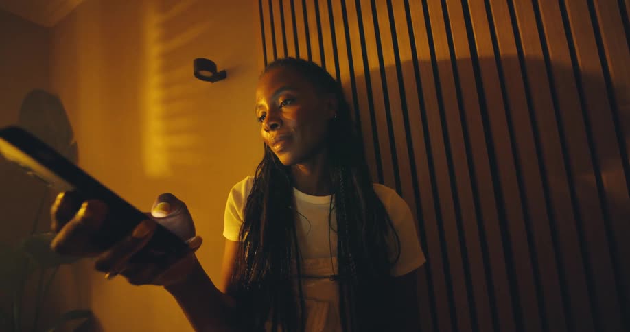 Woman with braided hair looking at phone indoors with warm lighting and wooden slat wall
