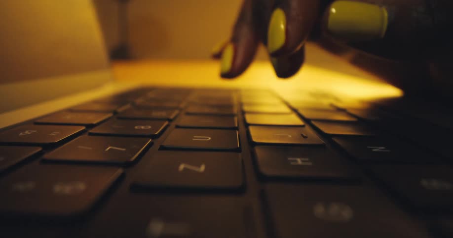 A dramatically lit closeup focuses on the dark keys of a laptop keyboard while a persons darkskinned hand with bright yellow nail polish is visible typing in the upper right illuminated by a warm golden light coming from the background