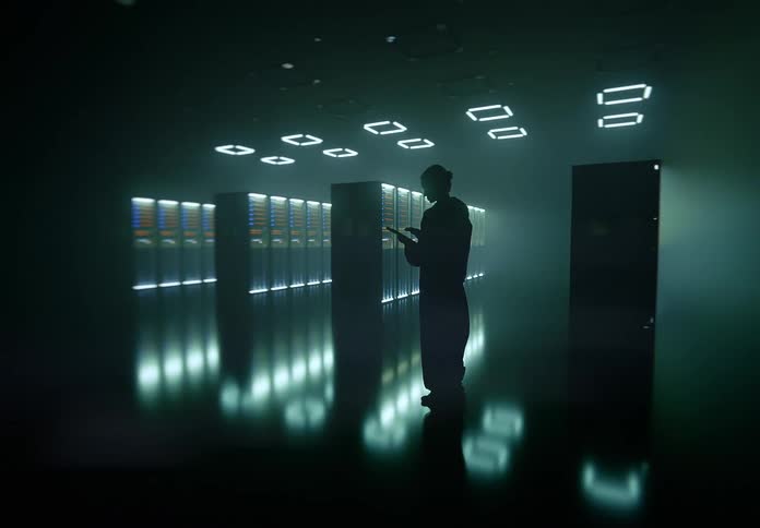 A lone figure stands in a shadowed server room in a data center before a grid of rectangular lights activates overhead casting a cool glow across the reflective floor