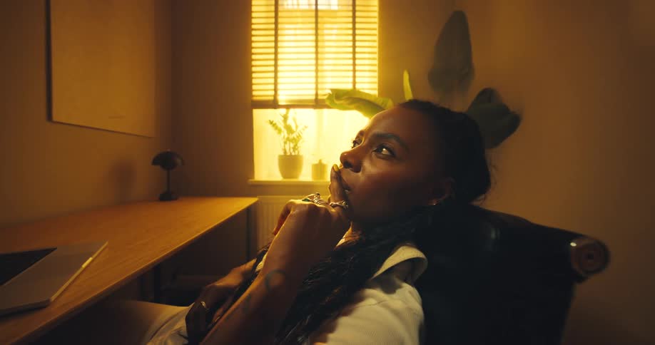Young woman sitting at a table in front of a laptop computer as she leans back in her chair thinking