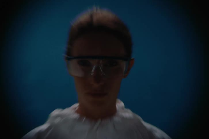A woman wearing safety goggles and a protective suit stands in dramatic blue lighting