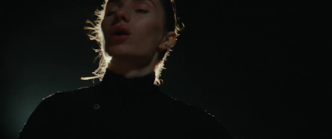 A woman with fair skin and her hair pulled back wearing a dark turtleneck intensely illuminated from behind by a single bright light that highlights her profile and hairline against a dark black background