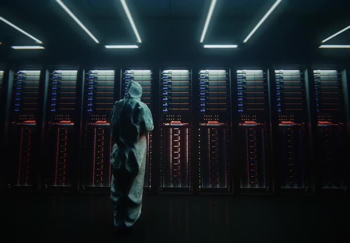 Beginning with a closeup of cables the view rapidly pulls back to reveal a lone figure in a hazmat suit facing a vast wall of blinking servers in a data center