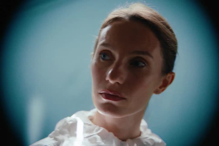 A young woman dressed in a white coat stands inside a room while a device scans her