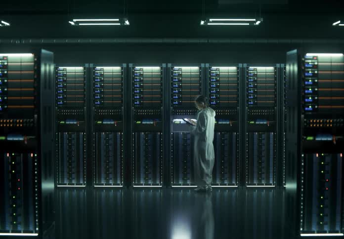 A person in a white protective suit inspects tall glowing server racks in a dark data center