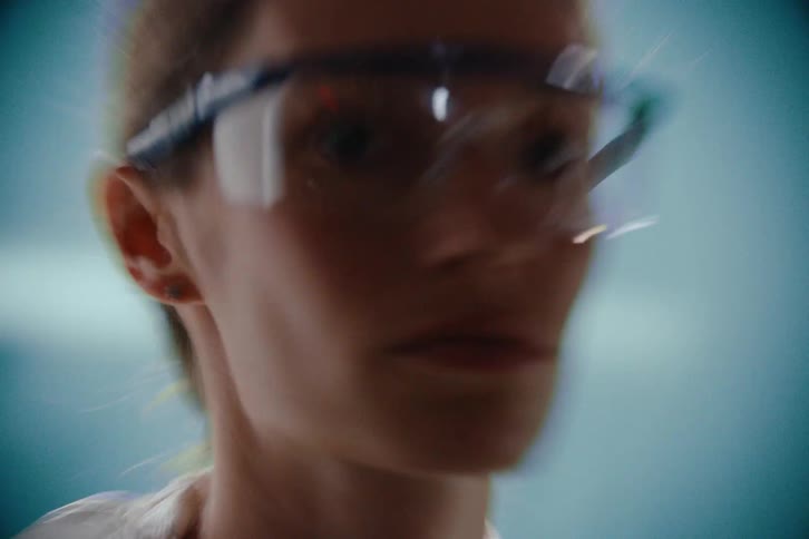 A young woman dressed in laboratory clothing and wearing goggles spins left and right inside a room