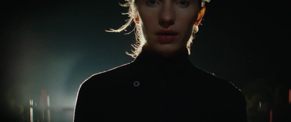 A woman breathing heavy with light hair and fair skin is dramatically backlit by a bright source creating a halo effect around her head as she looks directly forward in the dark