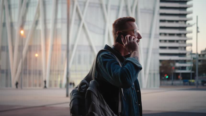 Man talks on the phone as he walks by buildings with a bag over his shoulder