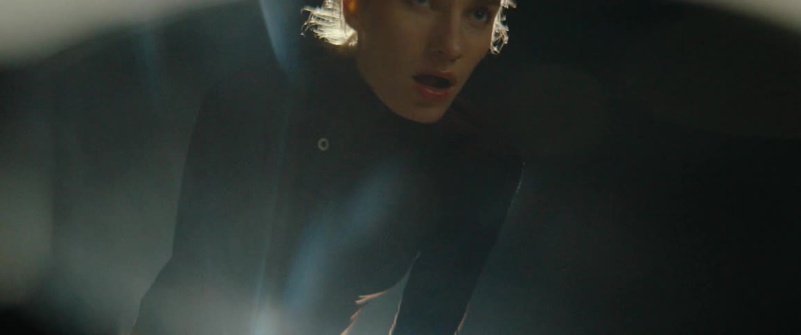The video shows a mediumclose up of a woman wearing a dark turtleneck filmed in low light with dramatic backlighting that highlights her hair and shoulders she is hunched over and appears to be breathing heavily possibly from exertion with her eyes w