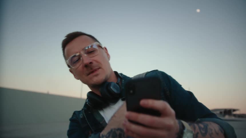 A man wearing a denim jacket clearrimmed glasses and headphones around his neck looks down and scrolls on his black smartphone while standing outdoors in the evening