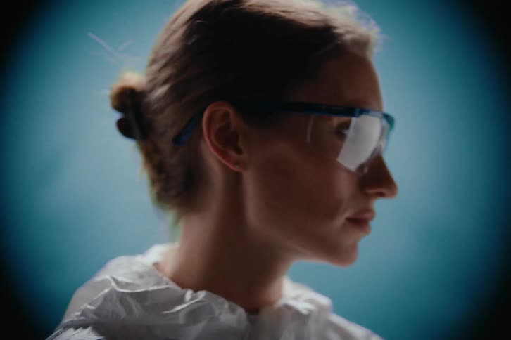 A closeup shot of a woman in a white protective suit and blueframed safety goggles whose focused and intense expression is illuminated by a strong central light source against a teal background as she turns her head