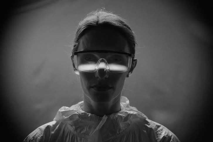 A young woman dressed in overalls and wearing goggles stands inside an elevator