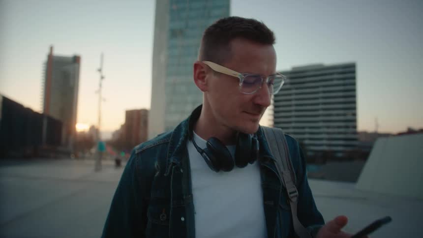 A man wearing glasses a denim jacket and a backpack with headphones hanging around his neck is looking down at a mobile phone he holds in an urban setting at dusk