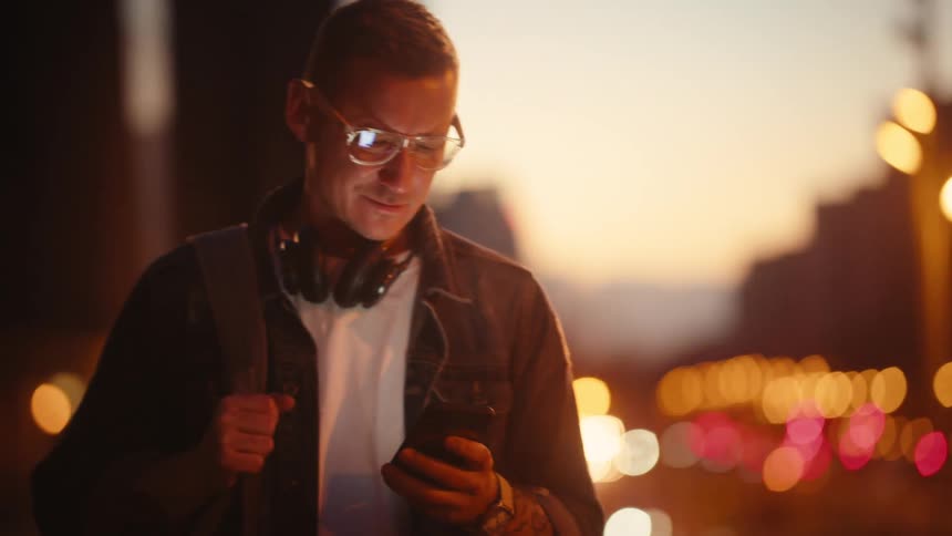 A man wearing glasses headphones around his neck and a backpack checks his cell phone while walking on a city street at twilight with the urban background illuminated by blurred lights