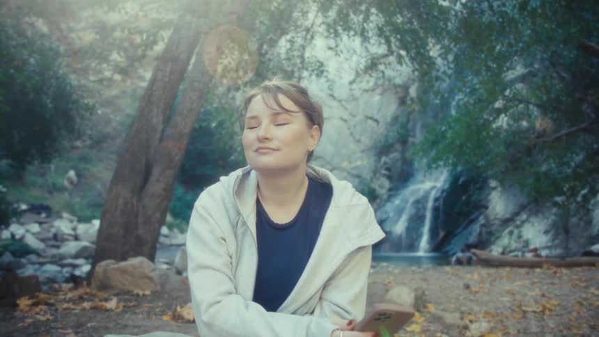 The video shows a woman sitting outdoors near a waterfall initially looking at her phone before setting it downshe then closes her eyes and takes a deep appreciative breath suggesting she is relaxing and enjoying the tranquil natural environment ar