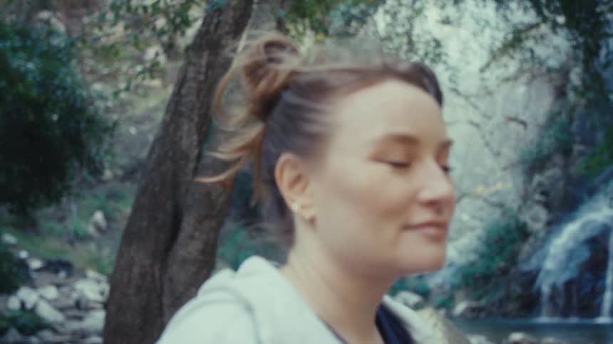 Woman with messy bun and light hoodie standing near waterfall in natural setting