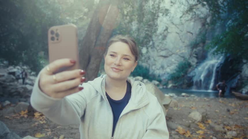 A woman sitting on a rock in a forest smiles as she lifts her phone to take a selfie