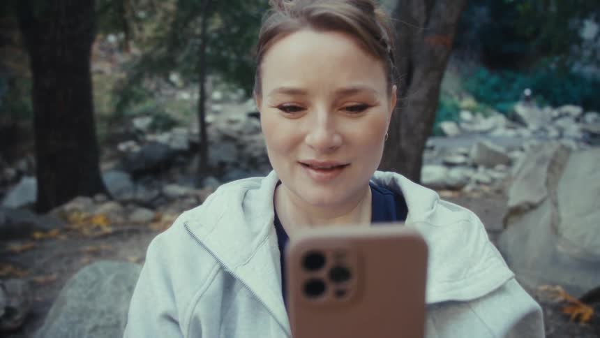 The video opens with a woman outdoors holding a large goldenbrown autumn leaf close to the camera obscuring her face she lowers the leaf to reveal herself and then immediately raises a modern smartphone to take a picture of herself or the surroundin