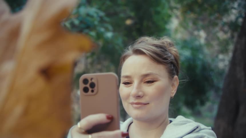 A woman with her hair pulled back holds up her smartphone to take a closeup picture of a large single brown and yellow autumn leaf in a park setting