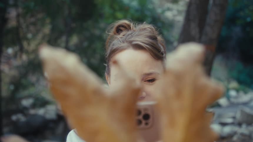 A smiling woman clicks a picture of a dry leaf with her phone in a forest