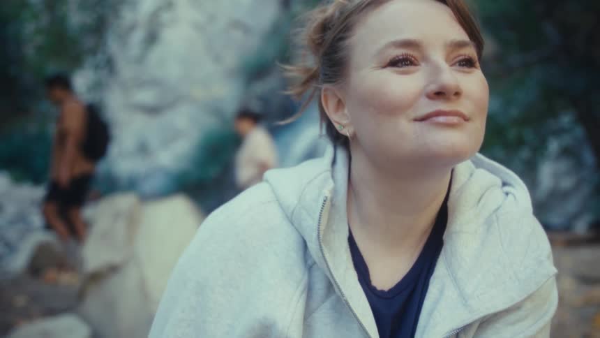 A young woman sits on a rock at midday wearing a hoodie and lost in deep thought during a hiking trip with a waterfall behind her and people setting up camp in the background
