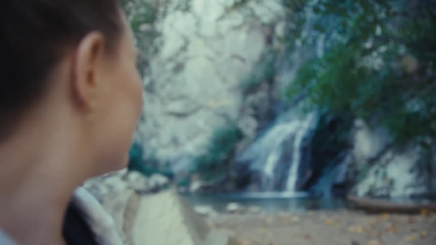 A woman with her hair pulled back and wearing a lightcolored hooded sweatshirt is captured in a medium closeup smiling gently while looking forward then behind at a visible waterfall