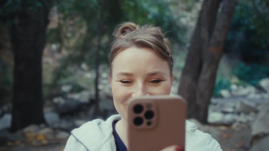 A lady holding a smartphone while standing in a forest area at dawn