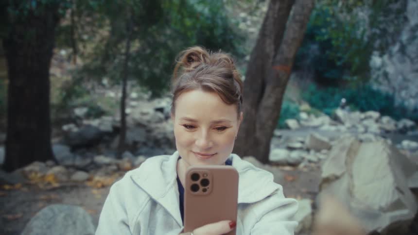 A woman with her hair pulled back holds up her smartphone to take a closeup picture of a large single brown and yellow autumn leaf in a park setting