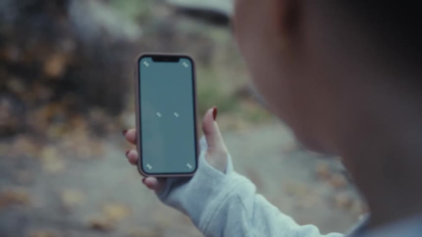 Smiling woman in light hoodie holding phone in front of blurred vegetation