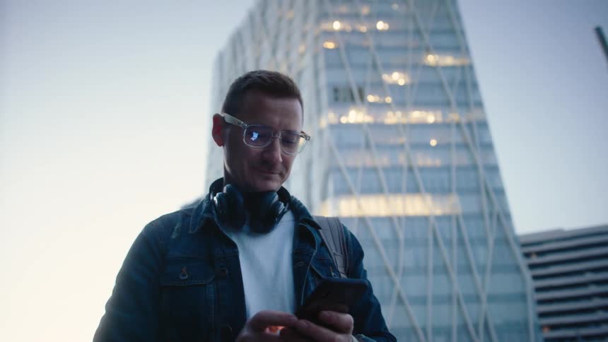 A man in glasses a denim jacket and headphones around his neck is standing in an urban setting he first holds his phone up to take a picture of a tall modern building and then looks down to interact with the device