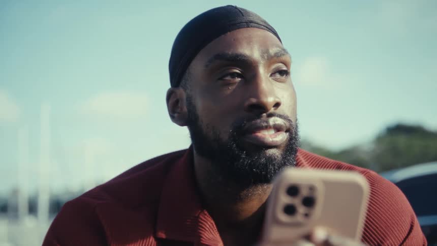 A man with a beard and a durag sits outdoors during the day holding a phone in front of his face then lowers the device slightly and looks up with a focused and serious expression