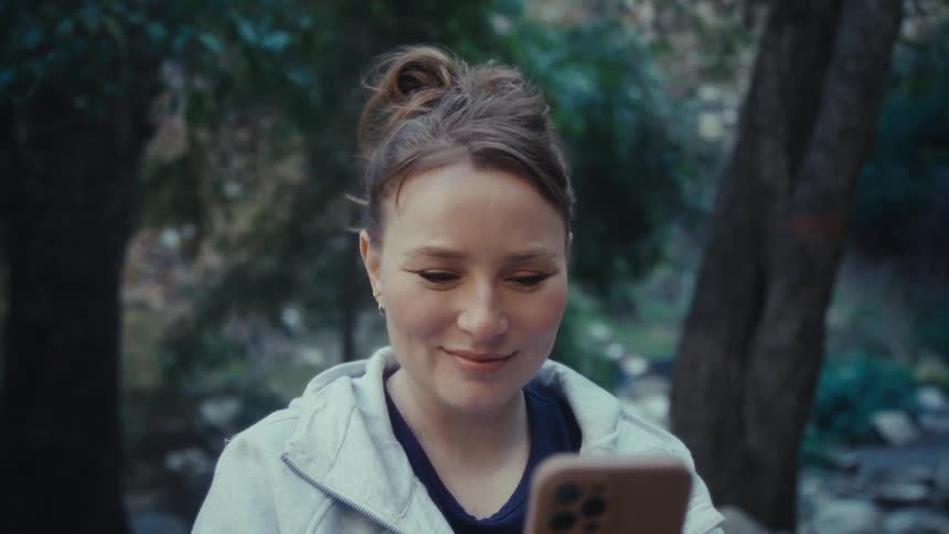 A closeup shot features a woman outdoors wearing a light gray hooded sweatshirt and a navy blue top with her hair pulled up she is holding a phone in a brown case and initially appears focused on the screen before looking up with a gentle genuine smi