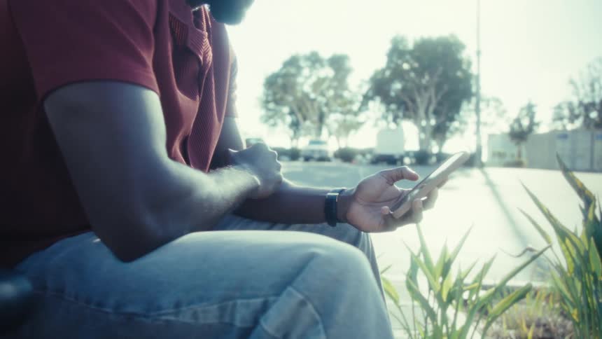 A man wearing a red shirt and a durag is sitting outdoors possibly in a park or by a road he is looking down at his phone and typing or scrolling