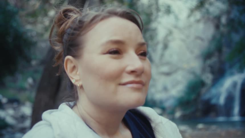A woman stands beside a waterfall and smiles while looking up