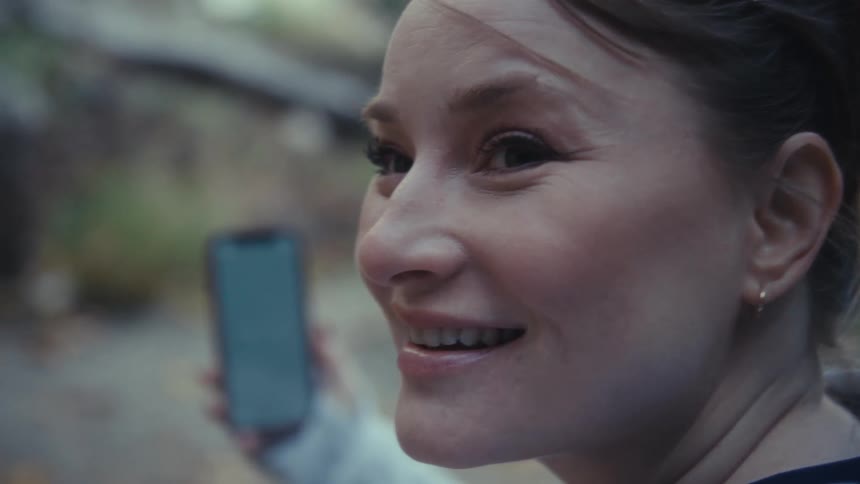 The video features a woman outdoors who initially holds up a smartphone with a blankgreenscreened display and smiles brightly offcamera as if recording a video or taking a picture the camera then focuses on the phone as she holds it steady with a