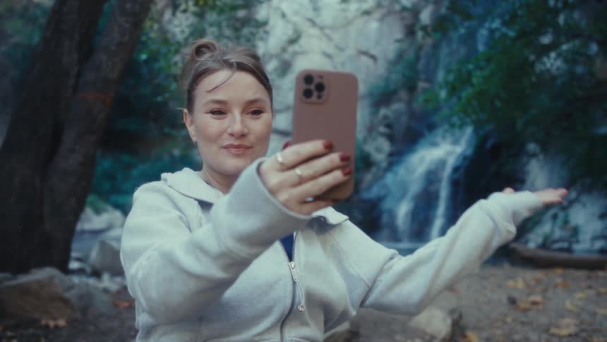 Woman smiles as she holds out her phone in conversation and looks at the waterfall