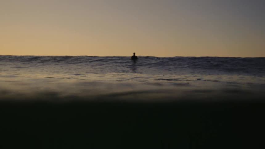 A man sits on a surfboard floating on the ocean at sunset as gentle waves create rippling effects across the water