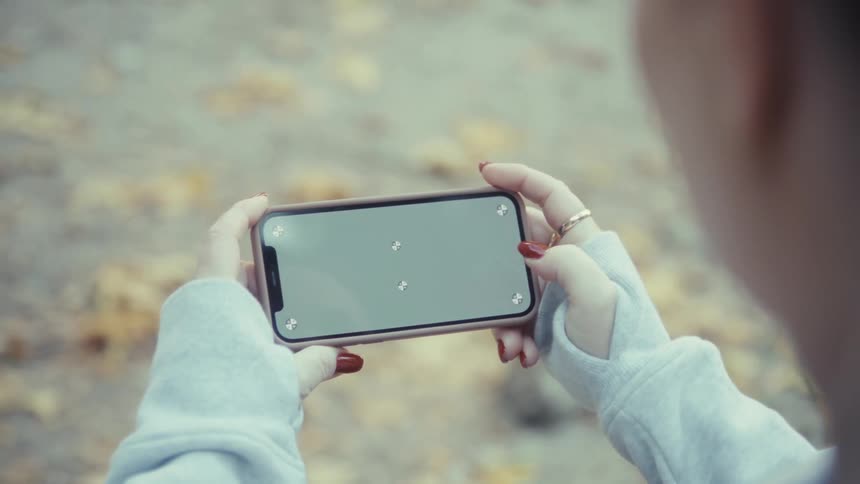 Woman outdoors tapping on the screen of a smartphone while sitting down next to dry leaves during day time