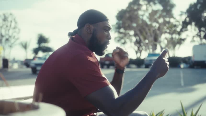 A sideprofile shot shows a bearded man outdoors on a sunny day wearing a rustcolored polo shirt and a dark durag actively using a mobile phone he appears engaged in a conversation or watching content gesticulating with his right hand as he holds the