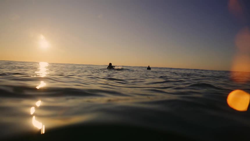 Two silhouetted surfers paddle out on choppy ocean waves under a vibrant sunset sky