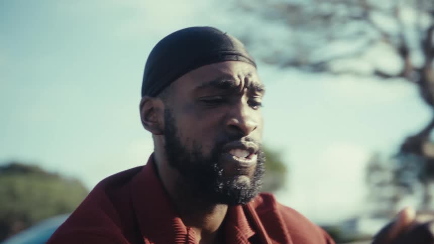 The video begins with a man wearing a red shirt and a durag sitting on an outdoor bench looking down at his phone and speaking intently as if on a video callthe camera quickly zooms in to a tight closeup of his face where his expression conveys dee